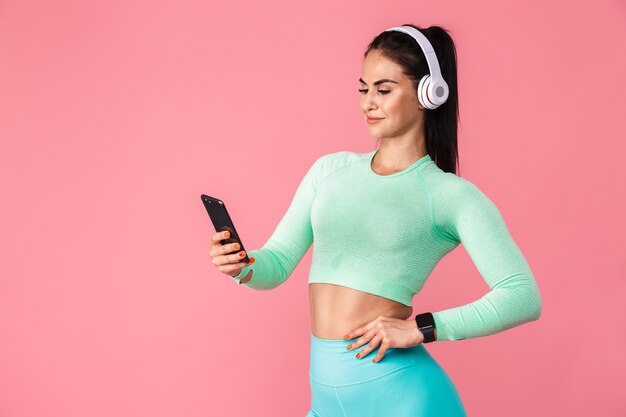 Portret van vrolijke blij mooie jonge sport fitness vrouw geïsoleerd over roze muur muur met behulp van mobiele telefoon luisteren muziek met koptelefoon.