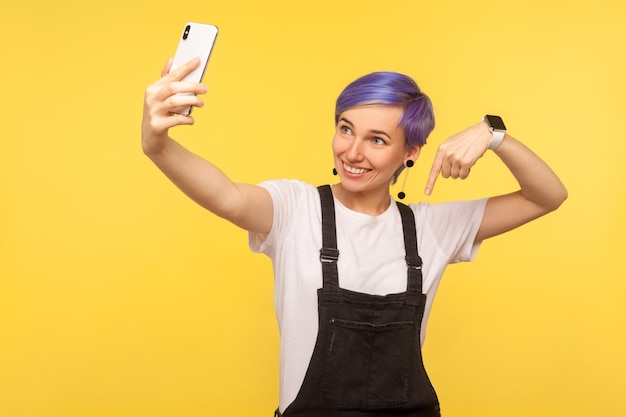 Portret van vrolijke blogger, hipster meisje met violet kort haar in overall met mobiele telefoon die videogesprek voert of blog online doet en naar beneden wijst, geïsoleerd op gele achtergrond studio opname