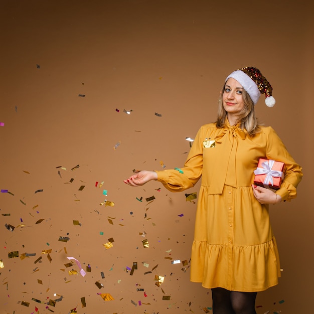 Portret van vrolijke blonde blanke vrouw in sprankelende kerstmuts en gele jurk gooien verpakt kerstcadeau met vallende confetti