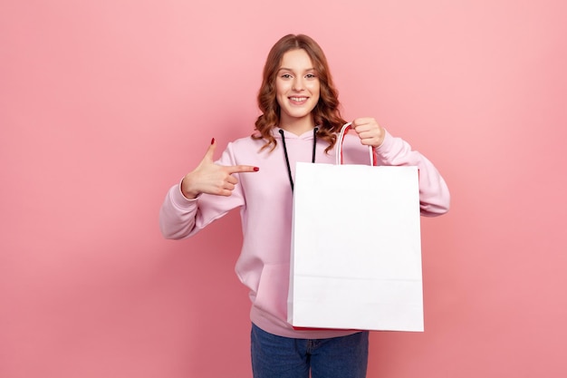 Portret van vrolijke brunette tienermeisje in hoodie wijzende vinger naar papieren zakken in de hand, kijkend met een brede glimlach, winkelen en verkoop. indoor studio-opname geïsoleerd op roze background