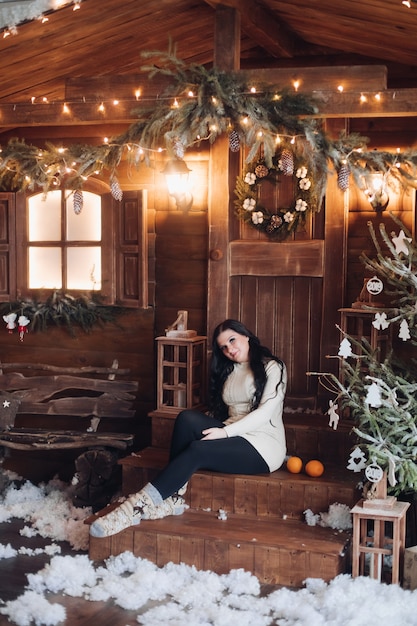 Portret van vrolijke en positieve vrouw met lang donker haar in trui, jeans en warme sokken met ingepakt cadeau voor Kerstmis zittend onder versierde kerstboom en sneeuwval