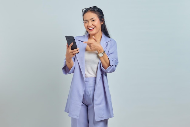 Portret van vrolijke jonge Aziatische vrouw die met de vingers wijst naar de mobiele telefoon die op een witte achtergrond wordt geïsoleerd