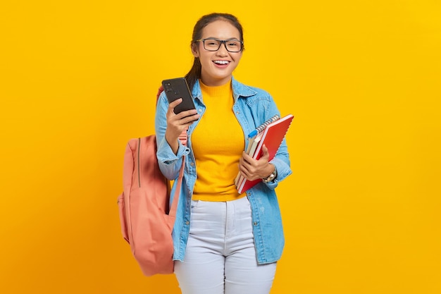 Portret van vrolijke jonge Aziatische vrouw student in denim kleding bril met rugzak met mobiele telefoon en boeken geïsoleerd op gele achtergrond Onderwijs in middelbare school universiteit college concept