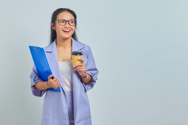 Portret van vrolijke jonge Aziatische zakenvrouw die klembord vasthoudt en afhaalkoffie drinkt geïsoleerd op paarse achtergrond