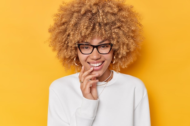 Portret van vrolijke jonge krullende vrouw met blije uitdrukking gekleed in casual witte trui glimlacht gelukkig draagt transparante bril poses tegen levendige gele achtergrond. Menselijke emoties concept