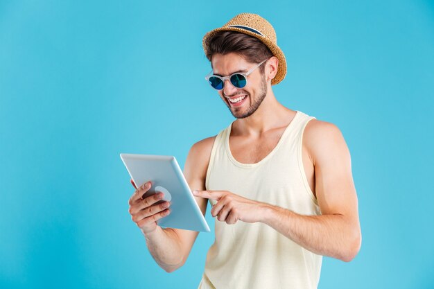 Portret van vrolijke jonge man in hoed en zonnebril met tablet over blauwe muur