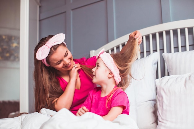 Portret van vrolijke jonge moeder en dochter die allebei roze t-shirts dragen die op bed in moderne slaapkamer zitten Vrouw en meisje in stijlvolle haarbanden onder kussens op bed Moeder die dochterhaar borstelt
