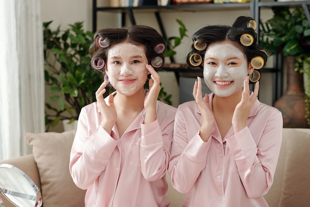 Portret van vrolijke jonge mooie vrouwen die nieuwe hydraterende en zuiverende gezichtsmaskers testen, ze zitten op de bank in zijden pyjama's en kijken naar de camera