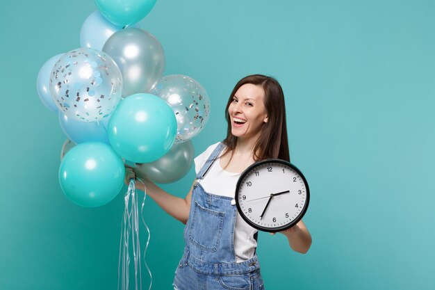 Portret van vrolijke jonge vrouw in denim kleding die ronde klok vasthoudt en viert met kleurrijke luchtballonnen geïsoleerd op blauwe turkooizen achtergrond. Vakantie verjaardagsfeestje, mensen emoties concept.