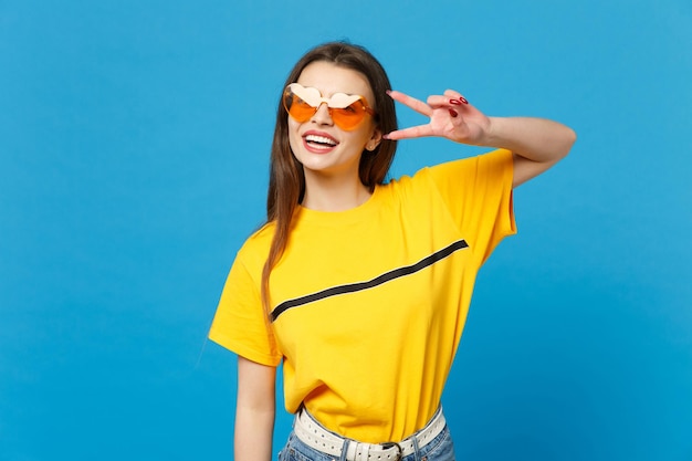 Portret van vrolijke jonge vrouw in levendige vrijetijdskleding, hartbril met overwinningsteken op zoek naar camera geïsoleerd op blauwe muurachtergrond in studio. Mensen levensstijl concept. Bespotten kopie ruimte.