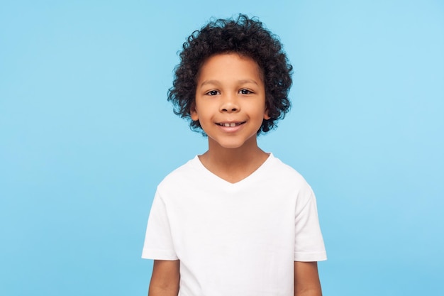 Portret van vrolijke kleine jongen met krullend haar in T-shirt glimlachend grappig en zorgeloos weergegeven: twee voortanden gezond gelukkig kind positieve emoties binnen studio opname geïsoleerd op blauwe achtergrond