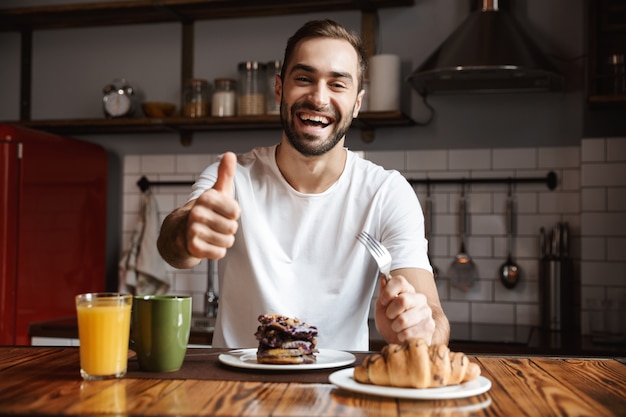 Portret van vrolijke man 30s croissants eten en sap drinken tijdens het ontbijt in stijlvolle keuken thuis