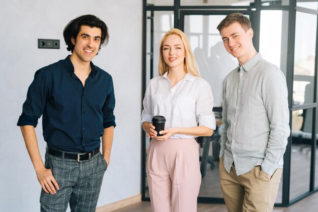 Portret van vrolijke mannelijke en vrouwelijke ondernemers glimlachend kijkend naar de camera die in een moderne coworking-ruimte staat Drie zakenmensen die een koffiepauze nemen