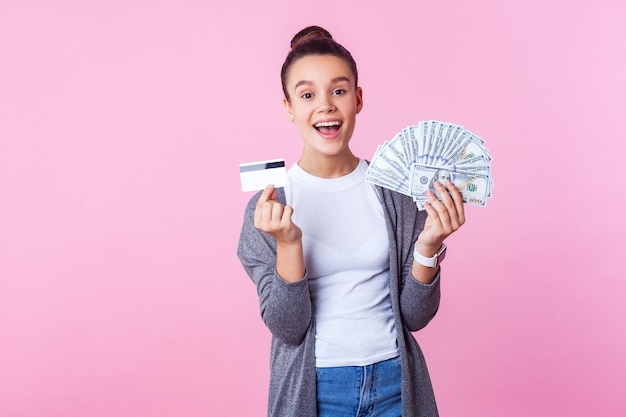 Portret van vrolijke opgewonden brunette tienermeisje met knot kapsel in casual kleding met dollarbiljetten en creditcard, kijkend met verbaasde vrolijke uitdrukking. studio-opname, roze achtergrond