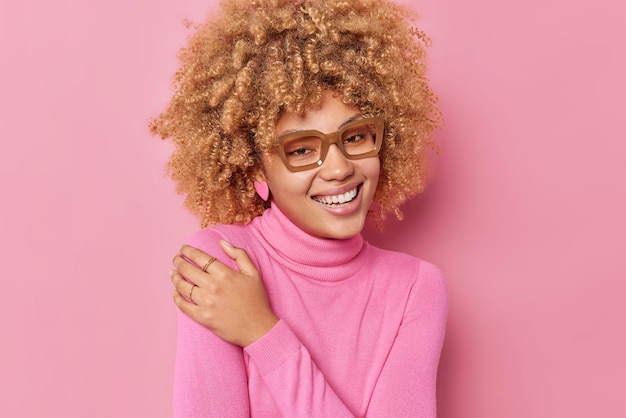 Portret van vrolijke oprechte vrouw met krullend haar houdt hand op schouder glimlacht breed toont witte tanden draagt bril en coltrui geïsoleerd over roze achtergrond. Gelukkig emoties concept