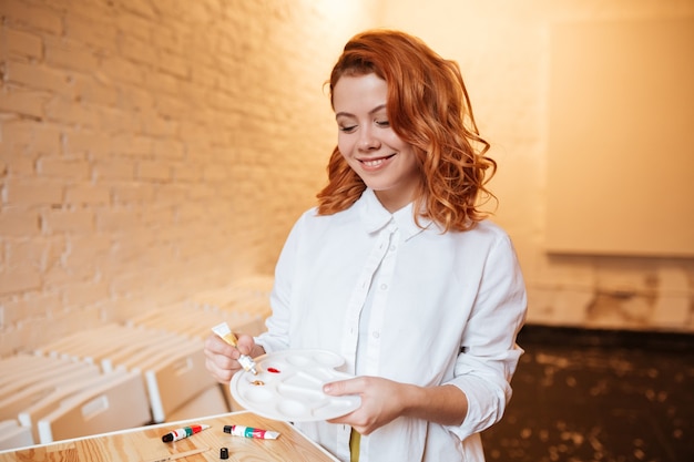 Portret van vrolijke roodharige jonge vrouwenschilder met olieverf en palet