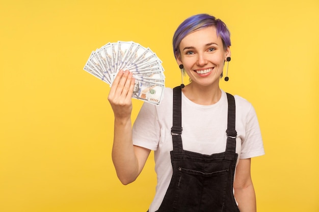 Portret van vrolijke stijlvolle hipster meisje met violet kort haar in denim overall fan van dollar biljetten tonen en kijken naar camera met brede glimlach geïsoleerd op gele achtergrond studio opname