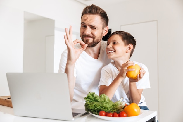 Portret van vrolijke vader en zoon die en laptop glimlachen gebruiken, voor het koken van maaltijd met groenten in keuken