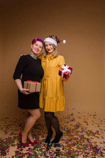 Foto portret van vrolijke vrouwen uitwisselen van kerstcadeaus en glimlachen. blonde draagt sprankelende kerstmuts en gele jurk