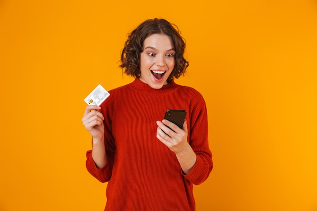 Portret van vrolijke vrouwen20s die sweater dragen die celtelefoon en creditcard met behulp van terwijl status geïsoleerd over geel