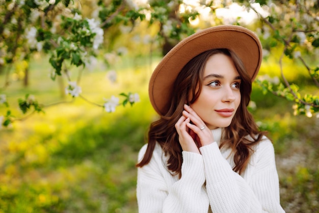 Foto portret van vrouw in hoed die poseert bij een bloeiende boom glimlachende jonge vrouw die geniet van de geur van bloemen