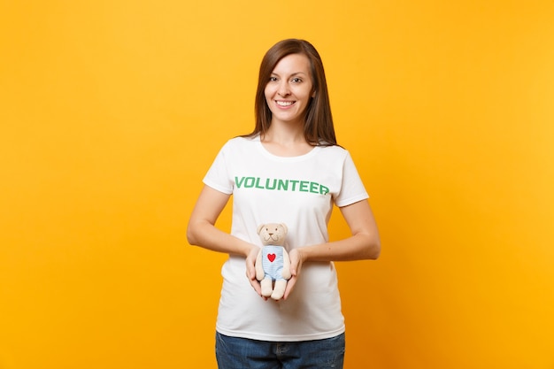 Portret van vrouw in wit t-shirt met geschreven inscriptie groene titel vrijwilliger houd teddybeer knuffel geïsoleerd op gele achtergrond. Vrijwillige gratis hulp, liefdadigheidswerkconcept.