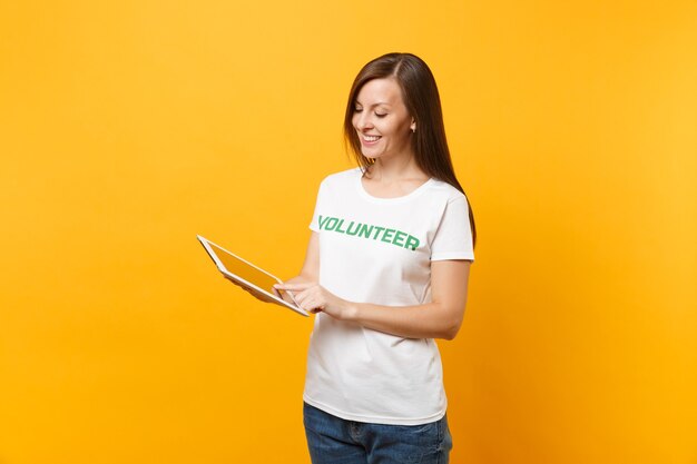 Portret van vrouw in wit t-shirt met geschreven inscriptie groene titel vrijwilliger met behulp van tablet pc-computer geïsoleerd op gele achtergrond. vrijwillige gratis hulp, liefdadigheidswerkconcept.