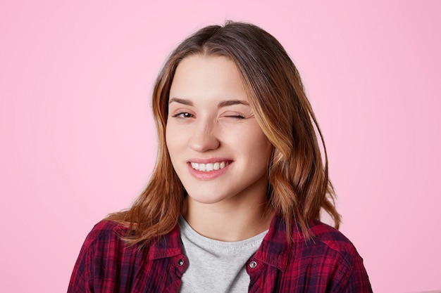 Portret van vrouw met vrolijke uitdrukking, knippert oog als flirt met knappe man, spreekt haar medeleven, gekleed in casual geruit overhemd, geïsoleerd op roze. Vrouw knipoogt binnen