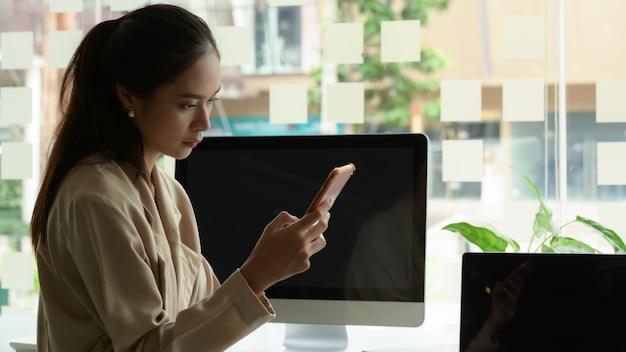 Portret van vrouwelijke beambte met behulp van smartphone zittend aan tafel met digitale apparaten in kantoorruimte