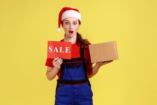 Portret van vrouwelijke koerier die pakket vasthoudt en verkoopbon toont bij bestellingen en levering met blauwe overalls en kerstman hoed Indoor studio opname geïsoleerd op gele achtergrond