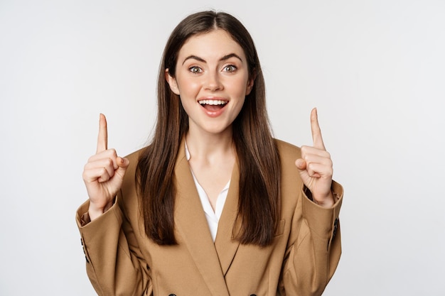 Portret van vrouwelijke ondernemer, zakelijke vrouw die met de vingers omhoog wijst, zelfverzekerd glimlacht, reclame toont, logo bovenaan, witte achtergrond