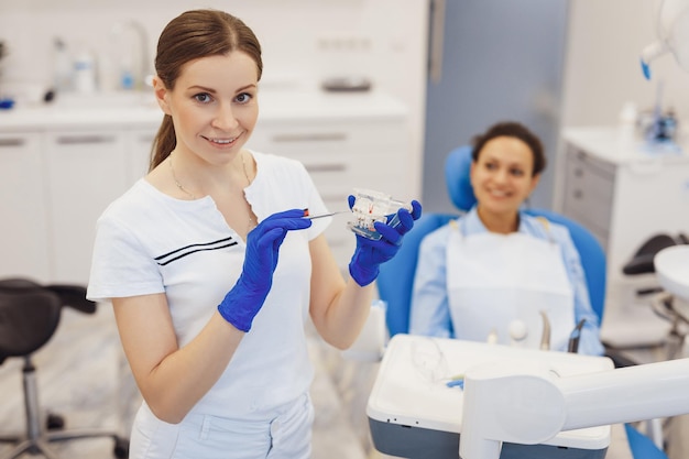 Portret van vrouwelijke orthodontist poseren op camera met kaken model in handen Vrouwelijke cliënt zittend in tandartsstoel op achtergrond