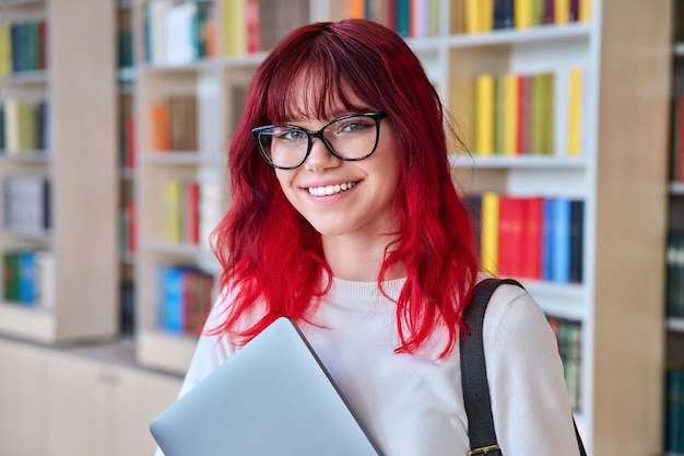 Portret van vrouwelijke student met laptop rugzak die naar de camera kijkt in de bibliotheek Mooi modieus meisje met gekleurd rood haar met een bril die glimlacht met tanden College universitair onderwijs jeugd