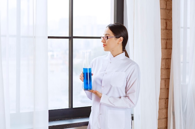 Portret van wetenschappervrouw in laboratoriumjas die chemische blauwe vloeistof houdt.