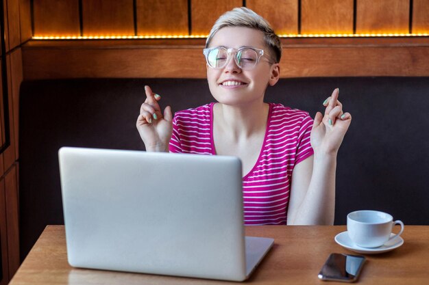 Foto portret van wishful aantrekkelijke jonge meisje freelancer in roze t-shirt zit in café en werkt aan laptop gesloten ogen en vraagt droom die uitkomt met een brede glimlach binnen levensstijl