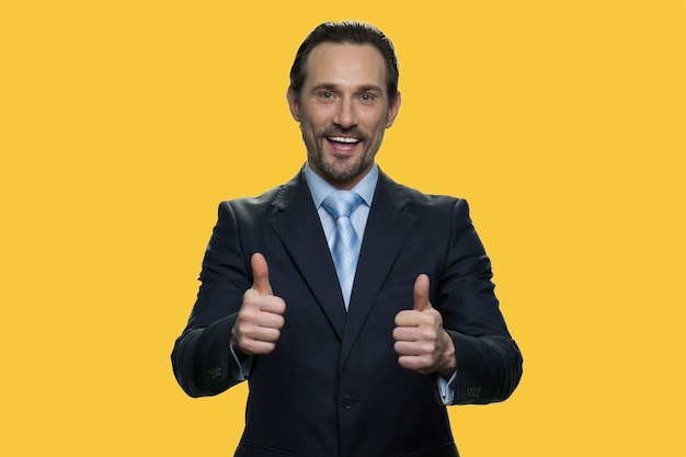 Portret van zakenman toont duimen. Zelfverzekerde positieve senior manager toont een gebaar als een teken van goedkeuring. Geïsoleerd op gele achtergrond.