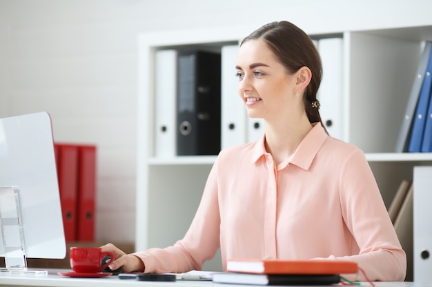 Portret van zakenvrouw. Hij zit in een kantoor aan een bureau en werkt op een bureaublad. Hij kijkt naar de monitor en glimlacht