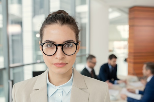 Portret van zelfverzekerde aantrekkelijke vrouwelijke advocaat in ronde brillen die tegen collega's staan die papieren op de achtergrond bespreken