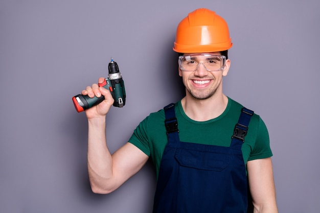 Portret van zelfverzekerde coole man reparateur houdt perforator klaar om plat appartement te vernieuwen draag blauwe overall groen t-shirt geïsoleerd over grijze kleur muur