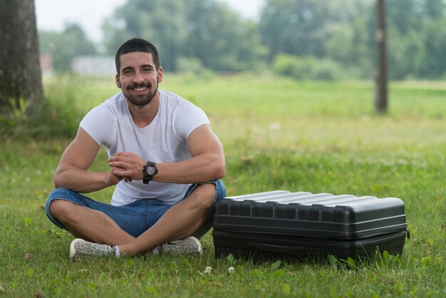 Portret van zelfverzekerde jonge ingenieur zittend op gras met koffer van drone