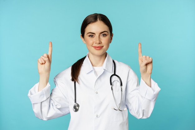 Portret van zelfverzekerde jonge vrouw arts medisch werker in jas wijzende vingers omhoog en glimlachend show...