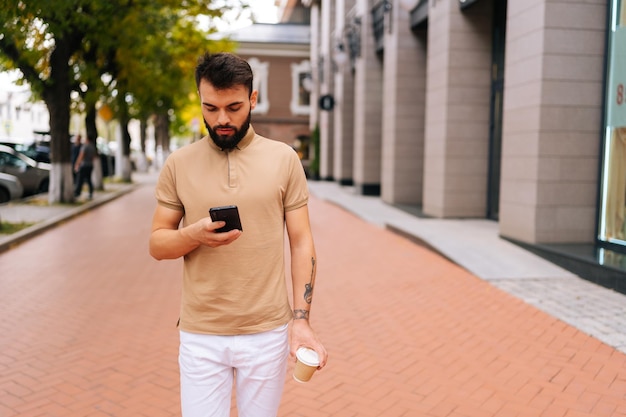 Portret van zelfverzekerde knappe jongeman met getatoeëerde hand die op straat loopt met smartphone en afhaalkoffie drinkt op zomerdag