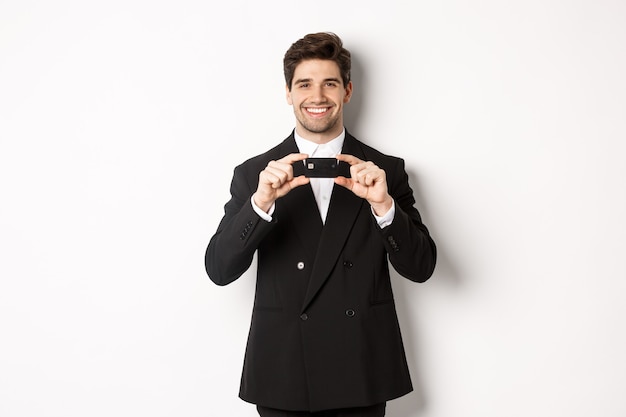Portret van zelfverzekerde knappe zakenman, glimlachend en creditcard tonend, staande in pak tegen een witte achtergrond.