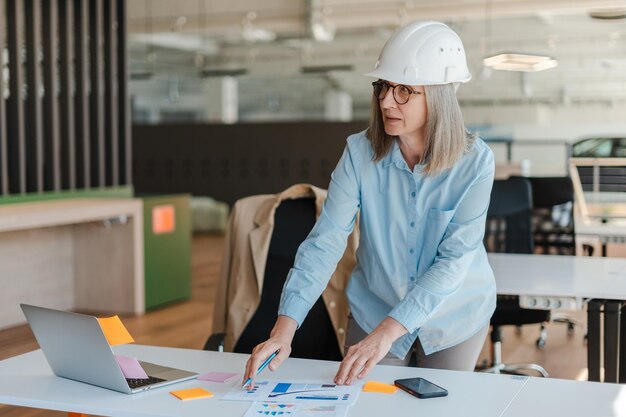 Portret van zelfverzekerde lachende senior vrouw architect-ingenieur met behulp van laptop werken in modern kantoor modern kantoor Succesvol bedrijfsconcept