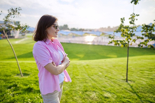 Portret van zelfverzekerde volwassen vrouw met gevouwen handen in shirtzonnebril, groen grasveld als achtergrondkopieerruimte, zonsondergang zomerdag