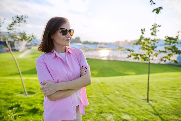 Portret van zelfverzekerde volwassen vrouw met gevouwen handen in shirtzonnebril, groen grasveld als achtergrondkopieerruimte, zonsondergang zomerdag