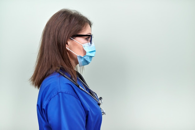 Portret van zelfverzekerde vrouwelijke arts met een stethoscoop medisch beschermend masker in blauw uniform op zoek in profiel op groene pastelachtergrond, kopieer ruimte