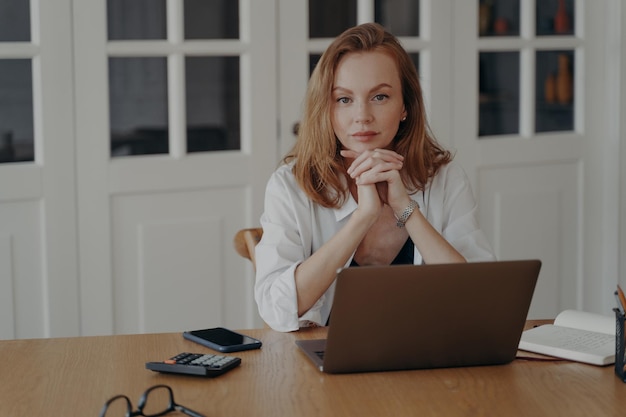 Portret van zelfverzekerde zakenvrouw die achter een laptop aan het bureau zit en naar de camera op de werkplek kijkt