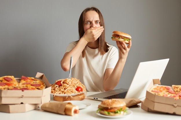 Portret van zieke overeten vrouw zittend aan tafel voor notebook geïsoleerd over grijze achtergrond sandwich vasthouden voelt slecht en misselijkheid bedekt mond met handpalm