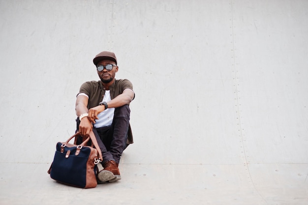 Portret van zittende stijlvolle Afro-Amerikaanse man draagt een zonnebril en pet met handtas buiten Street fashion zwarte man